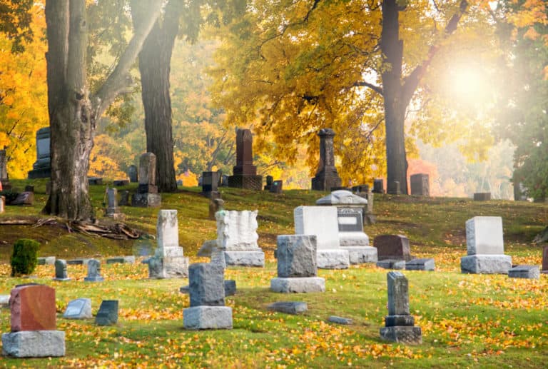 Cemetery in fall with upright gravemarkers and trees with yellow leaves.
