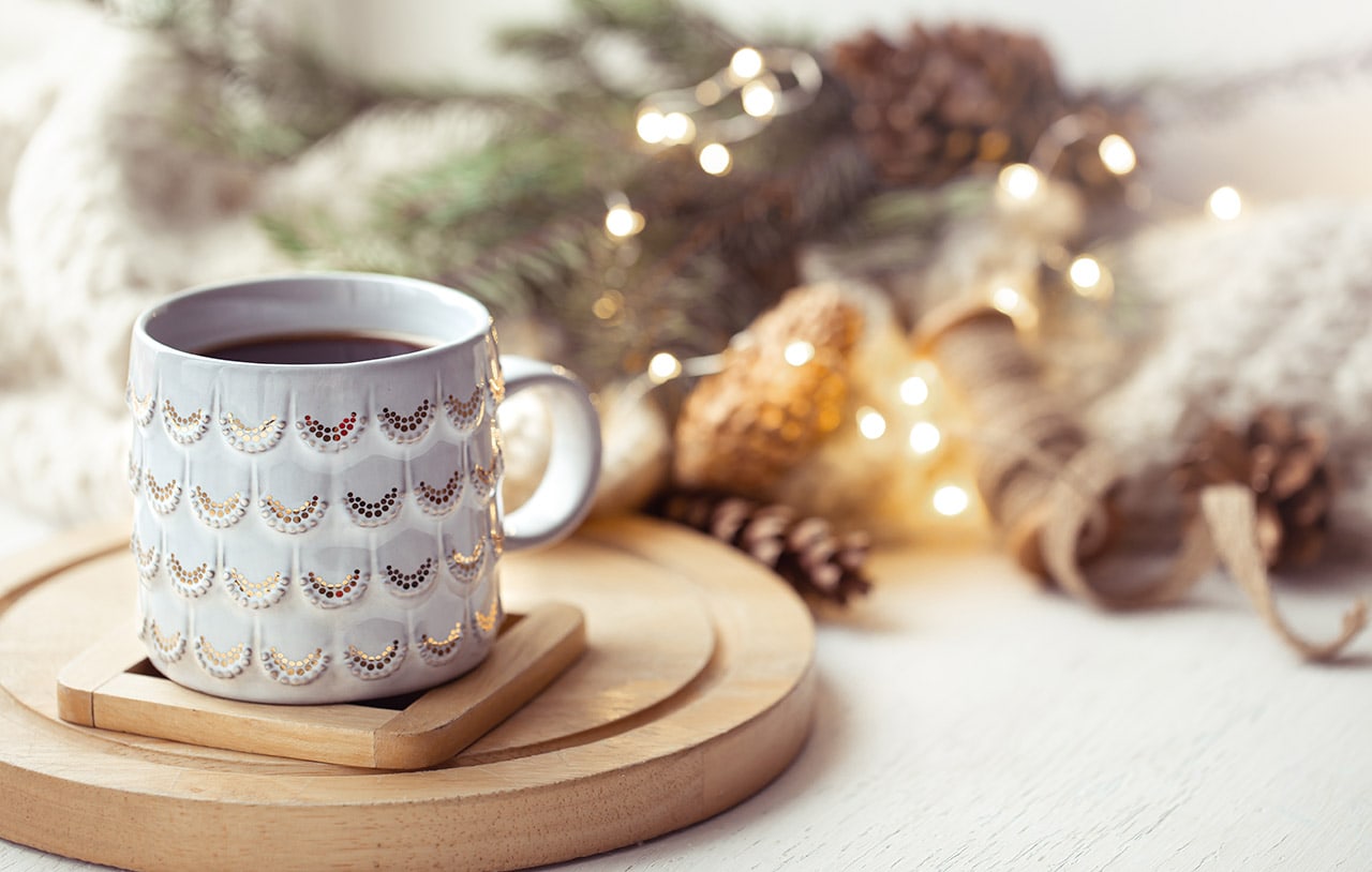 Image of a white holiday coffee mug with a blurred background of pinecones and lights.