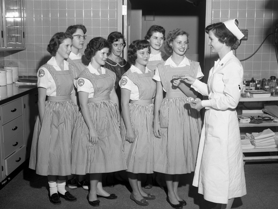 The Genealogy Blog Party Celebrates Women's History Month 2 Black and white image of seven young girls in candy striper uniforms being instructed by a woman in a nursing uniform.