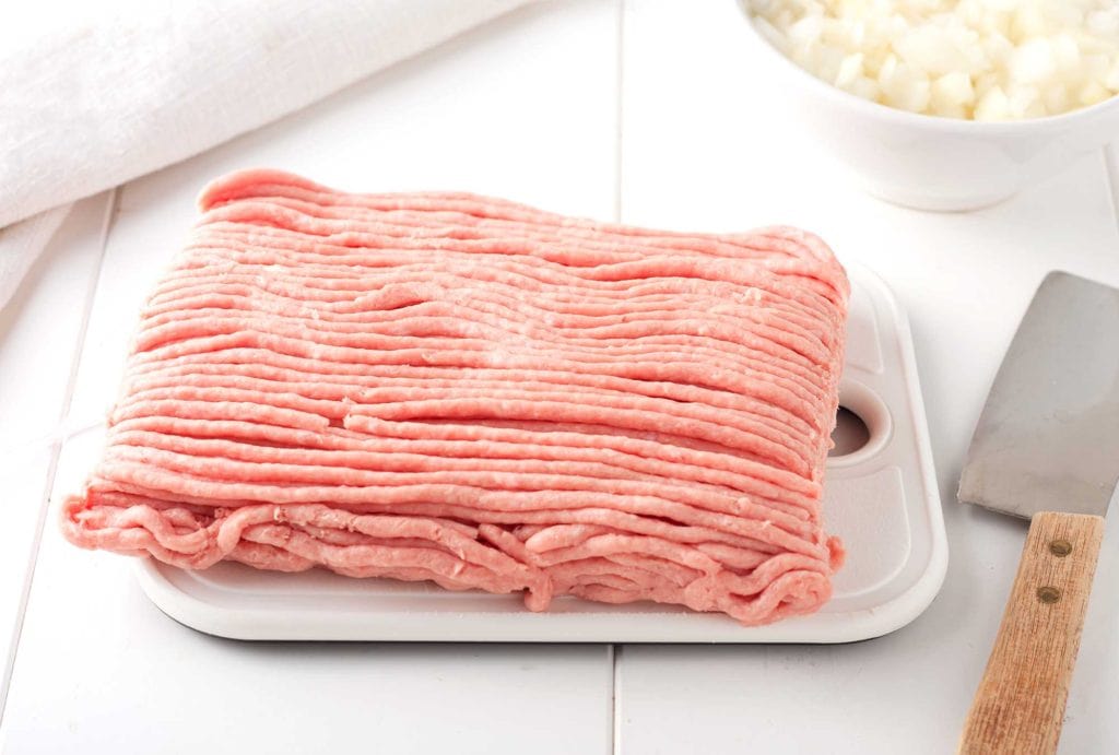 Turkey Meatloaf: An Update to a Mid-Century Classic Comfort Food 2 Image of uncut ground turkey on a white cutting board next to a bowl of white onions and a butcher's knife.