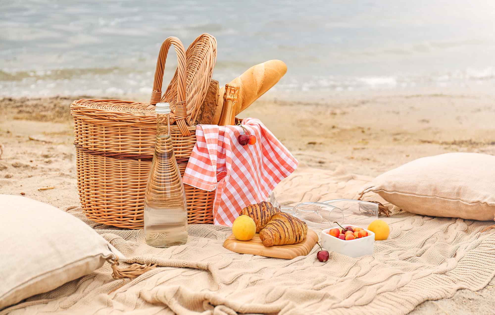 A picnic basket with croissants on a beige blanket with beige pillows by the ocean.