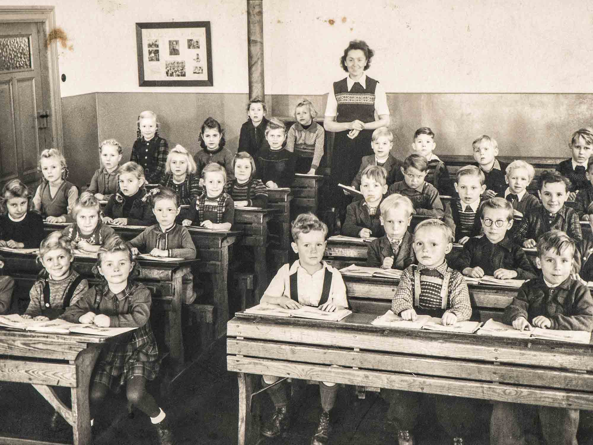 Sepia image of young students and a teacher in a c. 1940s school classroom.