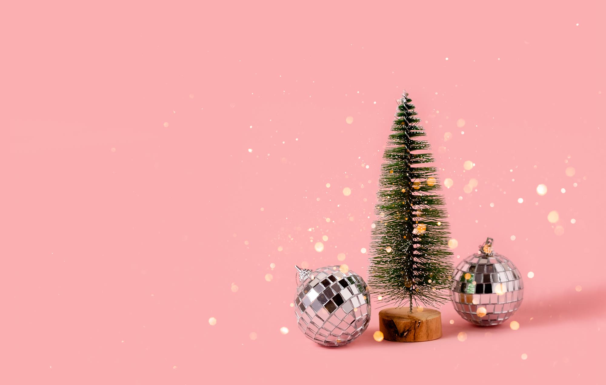 A toy fir tree next to two mirrored Christmas balls on a pink background with golden bokeh.