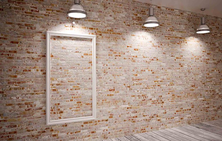 A red and white brick wall with an empty, white picture frame hanging on it and three overhead lights.