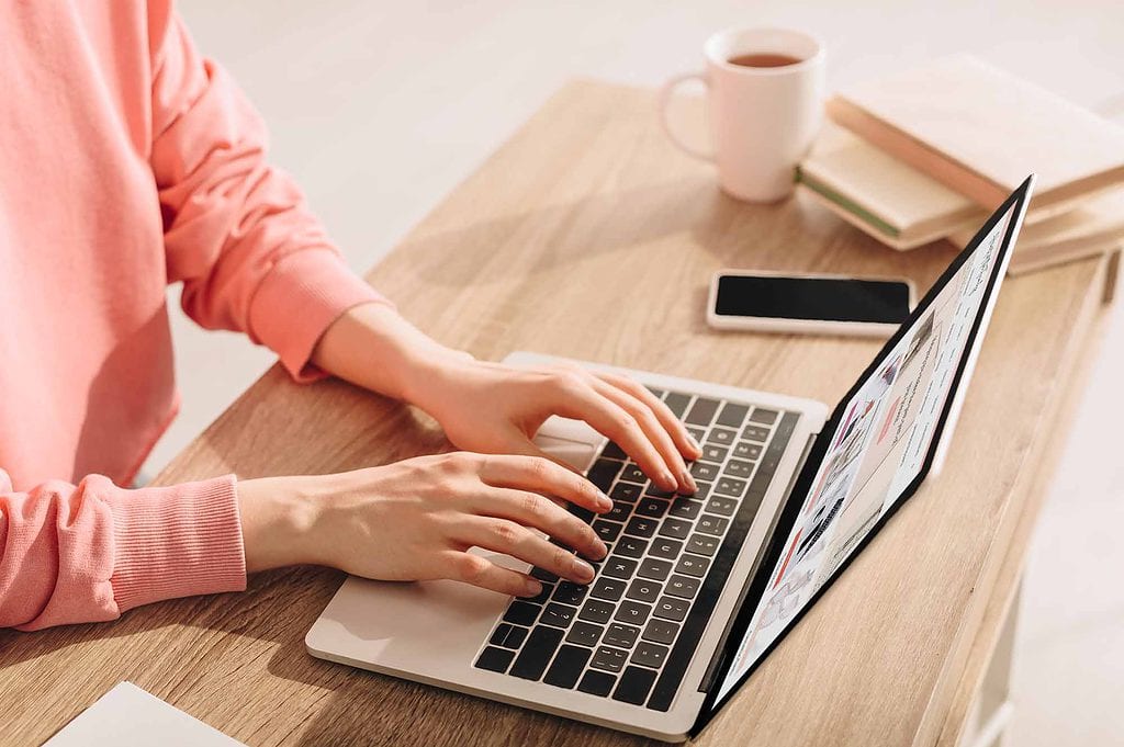 10 Best Strategies for Researching "Brick Wall" Ancestors 7 Hands of a woman in a pink sweatshirt typing on a laptop computer with a cellphone, books, and coffee on the desk.