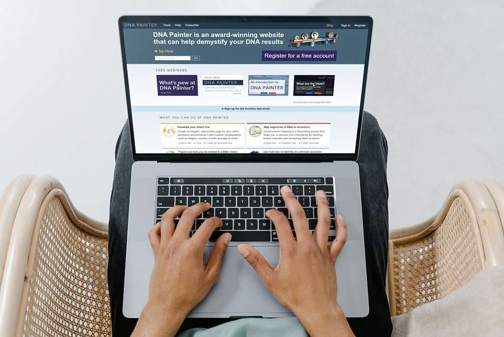4 Genetic Genealogy Tools I Use All the Time 3 Laptop computer displaying the DNA Painter website sitting on the lap of a man in a rattan chair with his hands on the keyboard.